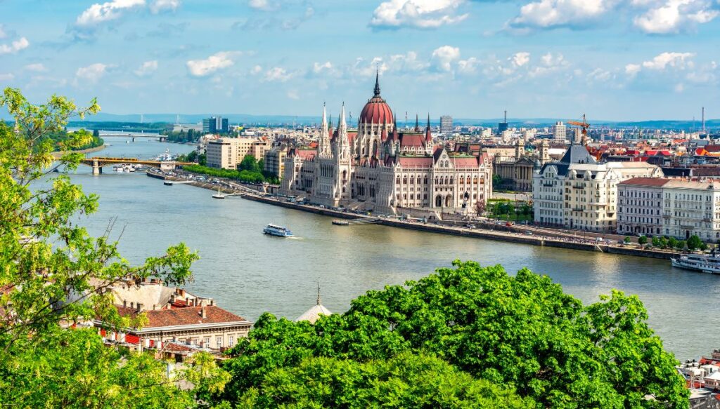 Panoramic view of the dome of the Parliament along the Danube River in Budapest with a boat sailing along the river on a sunny day.