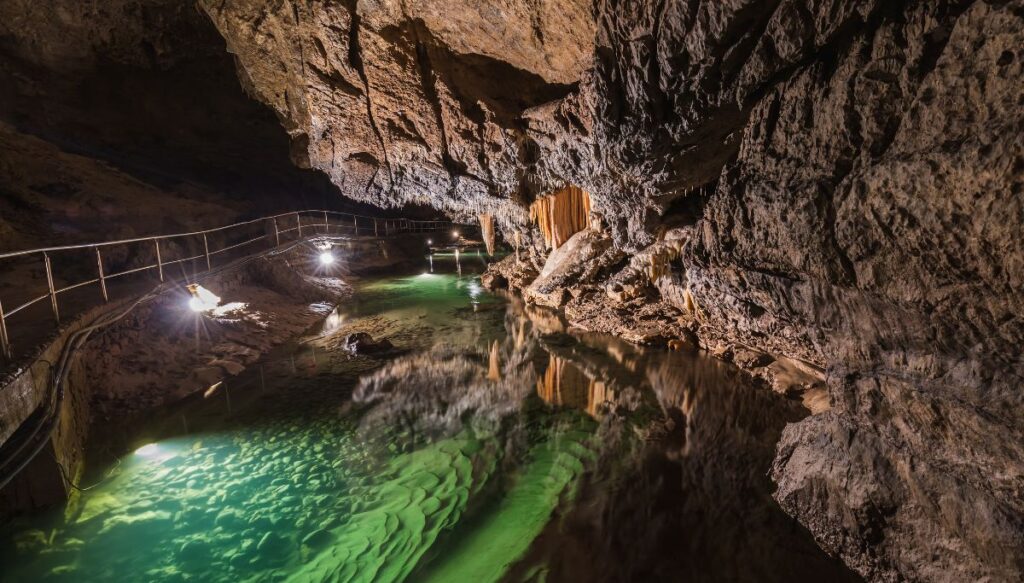 View of giant rock walls on the right and pool of water below it on the left with a railing and lights on the far left inside of the Cave of Liberty in Slovakia.