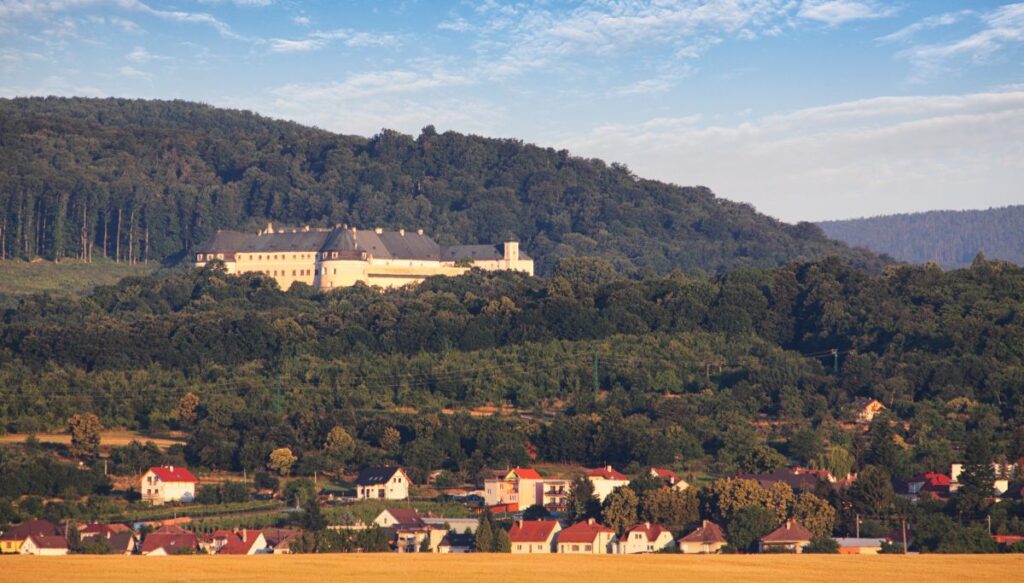 View of the white Červený Kameň Castle on the side of a hill surrounded by forests with a local Slovakian town sitting at the base of the castle.