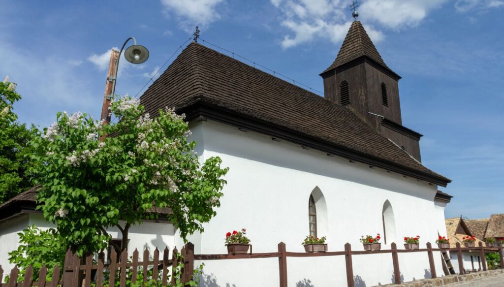 View of the white church in Hollókő with a dark brown roof and a steeple in the front on a sunny day in Hungary.