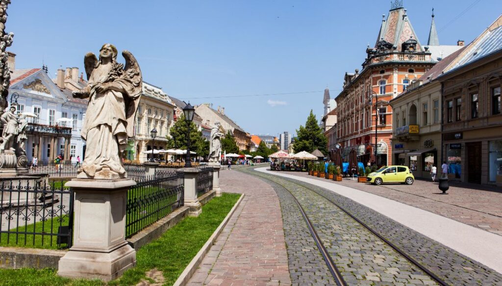View of an angel statue on the left, a train track through a stone path and historic buildings on the right in Košice, Slovakia.