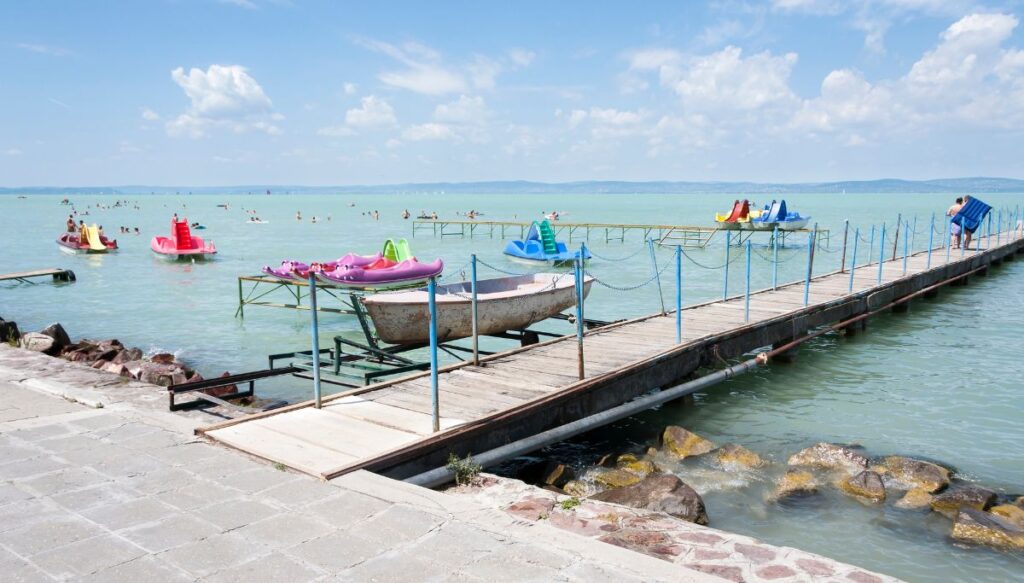 View of a wooden walkway extending into Lake Balaton with boats and slides in the water on a sunny day with rocks along the shore and a stone walkway in the foreground in Hungary.