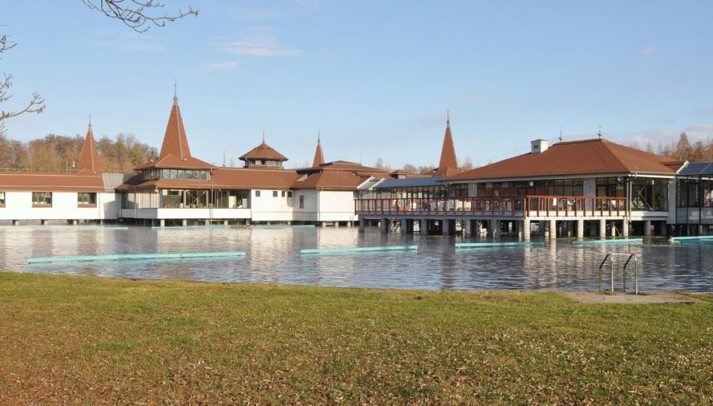 View of white buildings with brown roofs on Lake Heviz in Hungary with grass in the foreground and the lake in the background.