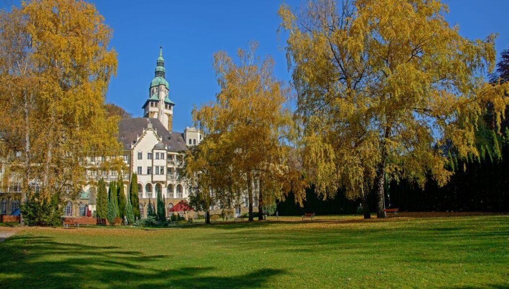 View of the Lillafüred Palace in white with teal roofs and a steeple in the background surrounded by yellow trees in Autumn.
