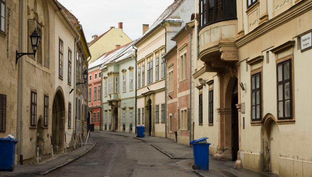 View of some of the pastel colored historic homes that line the streets of Sopron with blue trash cans in front of the buildings.
