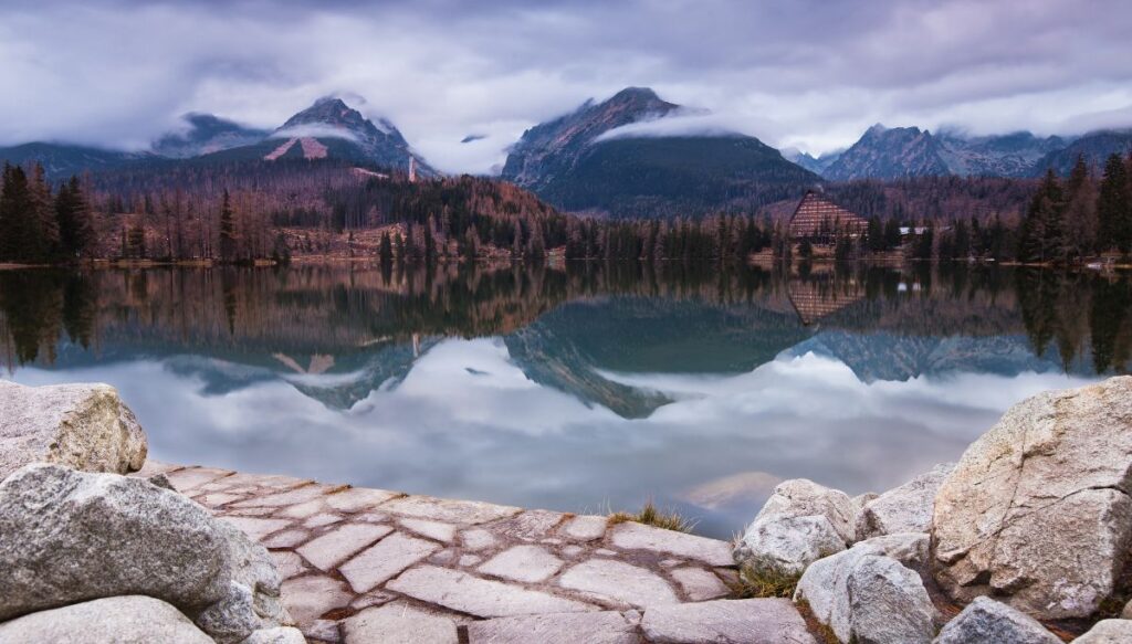View of a crystal blue lake with brown trees in the background and mountains hidden by clouds. You can also see white stones in the foreground at Štrbské Pleso.