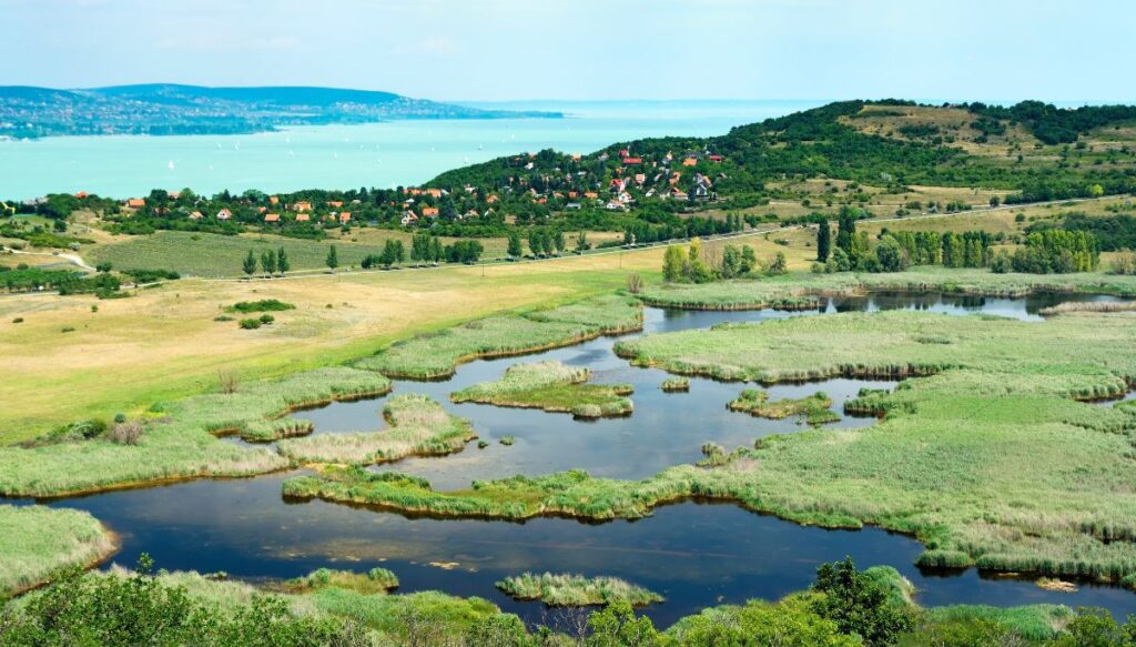 View of green fields inundated with water with green hills with houses by the lake in the background in Hungary.